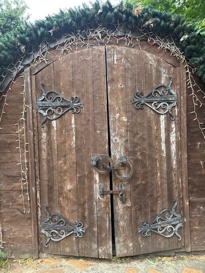 This is a close-up, eye-level, full-frame shot of a rustic, arched wooden double door. The doors are made of vertical planks with a weathered, distressed finish, showing significant wear and lighter colored patches. Each door is adorned with two ornate, dark metal strap hinges. In the center of the doors, there is a latch mechanism with two large, circular metal handles, and a small padlock secured to the latch.

Above the doors, a line of small, warm-toned string lights are draped, entwined with green artificial foliage that follows the curve of the arch. To the left and right of the doors, the wooden wall of the structure continues, also decorated with strands of the same string lights. The ground in front of the doors appears to be a pathway made of irregularly shaped stones and fallen leaves. The lighting suggests an overcast day. The location is identified as Yunost', Russia.