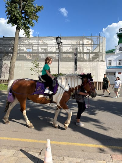 A young girl with brown hair, wearing a green t-shirt and black pants, is riding a brown and white pinto pony. The pony has a purple saddle pad with gold trim and a purple bridle. The pony is being led by a person wearing a gray shirt and black pants, with dark hair. They are walking on an asphalt road with a yellow line, next to a sidewalk made of gray paving stones. In the background, a large building is covered in scaffolding and a white tarp, with a prominent street lamp. To the right, two adults are walking on the sidewalk. The taller adult, a woman with long dark hair, wears a white t-shirt and khaki shorts. The shorter adult, a man with short dark hair, wears a dark green t-shirt and black shorts. The scene is set on a sunny day with a blue sky and scattered white clouds, in the city of Suzdal, Russia.