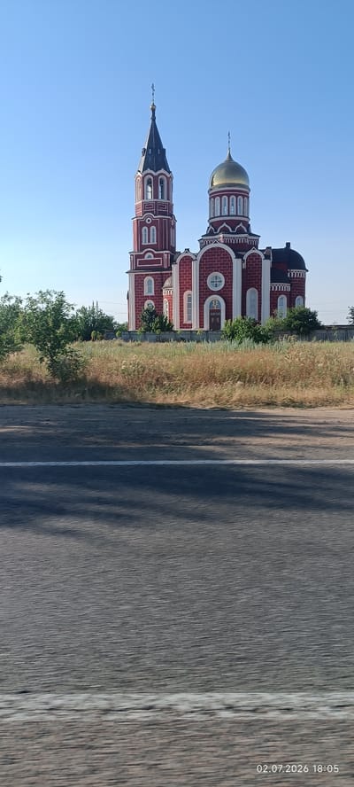 The image captures a bright, sunny day with a clear blue sky. Dominating the scene is a large, red and white orthodox church, featuring a tall bell tower with a dark spire topped by a cross, and a golden dome, also topped by a cross. The church exhibits intricate architectural details, including arched windows and decorative trim. In front of the church, a strip of dry, yellow grass and some green trees are visible. The foreground shows a paved road, with a white line marking the lane. The road appears slightly blurred, suggesting the photo was taken from a moving vehicle. Shadows of trees are cast on the road, indicating the sun is positioned to the side. The overall setting is rural, possibly on the outskirts of Velykyi Dalnyk, Ukraine, given the location context. There is no activity or event depicted, and no people are visible. The time is indicated by a timestamp in the bottom right corner: 02.07.2026 18:05. The atmosphere is serene and quiet, with the bright sunlight suggesting a warm, late afternoon.