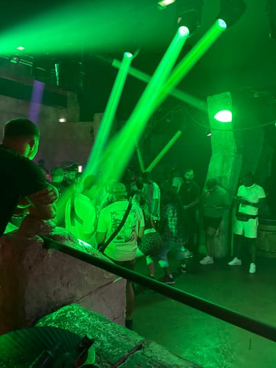 The image depicts an indoor event space, likely a club or concert venue, in Canet-en-Roussillon, France. Intense green laser lights dominate the scene, creating beams that cut through the dark atmosphere. Several people are visible, some standing and others in motion, suggesting a lively gathering. The environment features stone-like structures and what appear to be barrels, contributing to a rustic or possibly themed decor. One individual in the foreground, seen from behind, is wearing a dark t-shirt, while others in the background are dressed in casual attire, including t-shirts and shorts. The lighting conditions are low, with the green lasers being the primary source of illumination, casting a vibrant glow on the surroundings and the attendees. The overall impression is one of a nighttime entertainment event.