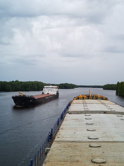 This is a wide shot of a river with two large barges moving on the water. The camera is positioned from the deck of a barge, looking forward. The barge in the foreground is empty, its deck covered with hatches. In the middle distance, another barge, black with a red stripe near the waterline and a white superstructure at the stern, is moving away from the viewer. The barge has the name "MEAH" written on its side in white Cyrillic letters. Both barges are navigating a wide river lined with dense green trees on both banks. The sky is overcast with thick, grey clouds, suggesting a cool or perhaps rainy day. The water is dark and calm, reflecting the muted light. The overall scene depicts a tranquil but functional moment on a waterway, likely in a rural or natural setting given the abundance of trees. The time of day is difficult to pinpoint precisely due to the overcast sky, but it appears to be daytime. The location is specified as Novokemskii, Russia. There is no indication of human activity beyond the operation of the barges themselves.