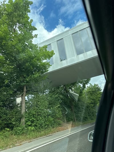 The image, taken from inside a vehicle looking out a window, captures a bright outdoor scene. A dark, angled portion of the car's interior frames the top and left edges, with noticeable reflections on the glass surface.

The prominent subject is a modern, multi-story building featuring a light-colored facade, likely white or light grey. It is characterized by horizontal rows of large, rectangular windows that reflect the blue sky and surrounding greenery.

In front of the building, and extending towards the foreground, is abundant vegetation. This includes numerous trees with full, verdant canopies and dense green bushes, alongside stretches of grass. A substantial tree trunk is visible in the mid-ground.

To the right, a section of an asphalt road is visible, defined by a distinct white lane marking and a grey curb or barrier. A car's side mirror is partially visible on the far right, confirming the perspective from within a vehicle.

Above the structures and foliage, a clear blue sky is dotted with fluffy white clouds, suggesting good weather conditions. The scene depicts a tranquil daytime setting, possibly in Zurzach, Switzerland. No specific activity or event is taking place, and no text is visible.