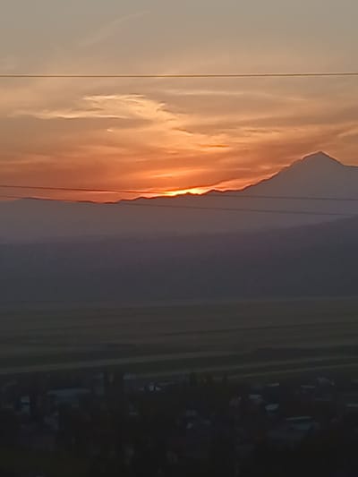 The image depicts a dramatic sunset over a mountainous landscape in Taran-Bazar, Kyrgyzstan. The sun, a brilliant orange orb, is setting behind a rugged mountain range, casting a warm, fiery glow on the clouds. The clouds are painted in shades of orange, pink, and gold, creating a breathtaking spectacle.
The foreground of the image is dominated by the sky and the mountains. The sky transitions from a light, pale orange near the horizon to a deeper purple and gray as it ascends. The mountains are silhouetted against the vibrant sky, their jagged peaks piercing the horizon. There are some faint, thin lines that appear to be power lines or perhaps the outline of distant structures.
To the right of the mountains, the landscape transitions to a darker, more subdued scene. This area appears to be a valley or a plain, with the details of buildings or terrain obscured by shadow and distance. The overall impression is one of natural beauty and grandeur, with the sunset highlighting the rugged terrain of Kyrgyzstan. The time of day is clearly sunset, and the weather appears clear, allowing for an unobstructed view of the celestial display. There is no visible text in the image.