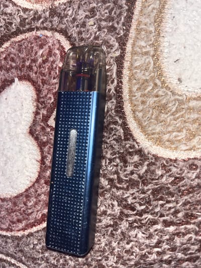 A blue vape pen with a clear cartridge is positioned vertically on a textured rug. The rug has a pattern of fuzzy fibers in shades of brown, white, and possibly some metallic threads. The vape pen has a textured, diamond-patterned body and a clear window showing a small amount of liquid. The top of the vape pen is clear and shows internal components, including colored plastic and metal. The lighting is dim, casting some shadows. There is no indication of the location being in Minyar, Russia.