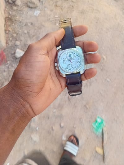 A dark-skinned right hand holds a silver-cased, square-shaped wristwatch with a skeletonized dial and a dark brown leather strap. The watch features visible internal mechanisms, a small detail at the 3 o'clock position, and a buckle on the strap. Below, a portion of a left foot wearing a black sandal with white stripes is visible on a light brown, unpaved ground. The ground surface consists of dirt and small stones, with scattered minor debris including a green plastic object and reddish fabric. The scene is outdoors in Dorayi, Nigeria.