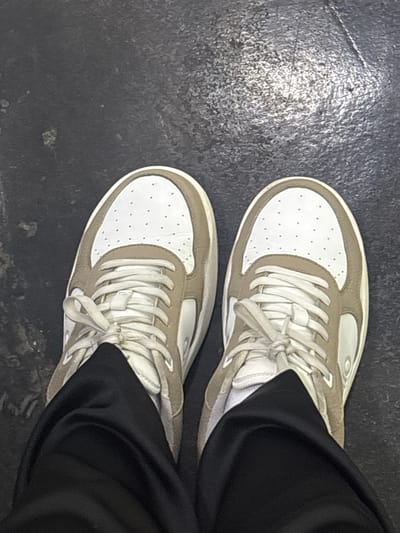 A top-down view of a pair of sneakers on a dark, possibly concrete floor. The sneakers are predominantly white with tan/beige accents around the edges and on the sides. The laces are white and tied. The right side of the frame is partially obscured by black fabric, likely pants, suggesting the perspective is from the person wearing the sneakers. The floor appears to be in an urban or indoor environment. No information indicates this image was taken in Buenavista, Mexico.