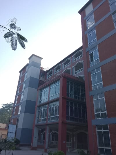 A low-angle shot captures several multi-story buildings. The rightmost building features a brick facade with vertically aligned windows and gray trim. A connecting structure with a red frame and large windows links it to another building with open-air balconies. To the left, a taller building with gray and blue panels also has windows. A tree branch is visible in the upper left corner against a light sky.