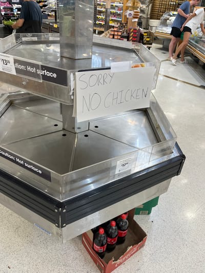 The image shows an empty display case in a supermarket in Middle Park, Australia. The case is made of stainless steel and has a black trim. It is empty, with the exception of a handwritten sign that reads "Sorry no chicken." There is a cardboard box with six bottles of soda lying on the floor next to the case. The case is labeled "Caution: Hot surface." There is a person standing at the back of the display case, out of focus. The floor is tiled in white and grey. The background is blurred, showing the shelves of a grocery store. The coordinates of the location are -27.556 latitude and 152.922 longitude.