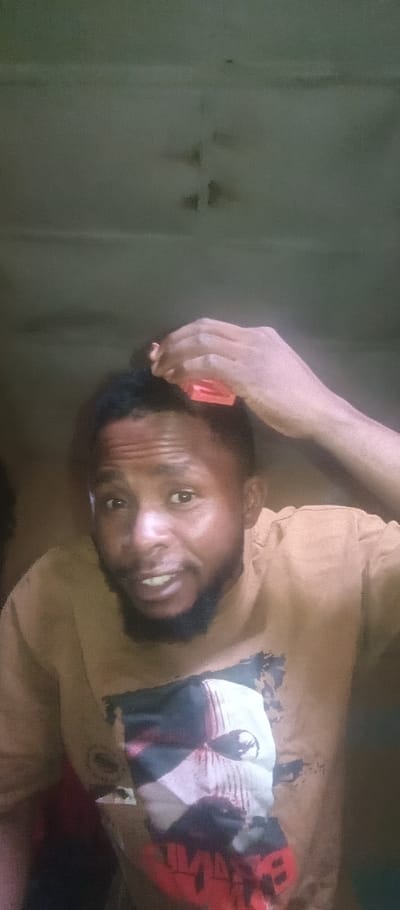 A man with dark skin, a beard, and short dark hair is holding a red comb near his head. He is wearing a t-shirt with a faded graphic design, possibly a face with some red elements. The background appears to be a dimly lit interior space with a plain ceiling. The photo's perspective suggests it was taken at a lower angle, looking up at the man. The overall image quality is somewhat grainy, suggesting low lighting conditions.