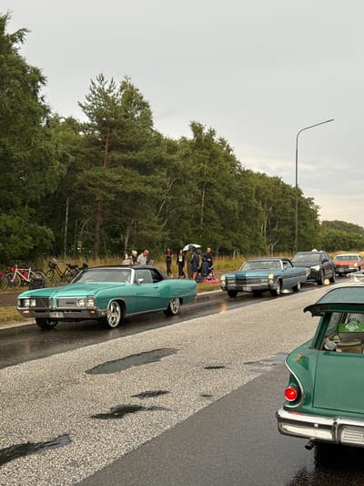 The image captures a lineup of classic American cars on a wet asphalt road, likely during an event in Falkenberg, Sweden. The sky is overcast, suggesting rain, which has left puddles on the road surface. A line of lush green trees borders the road on the left, with a power line visible in the distance on the right.
In the foreground, a vintage green convertible, possibly a Buick, is parked facing left. Its chrome details and custom wheels are prominent. Behind it, a light blue classic Cadillac is seen, followed by a dark SUV and an orange classic car. Further back, a green station wagon with its trunk open reveals some items, including a green box with "Carlsberg Hof" branding.
Several people are gathered on the left side of the road, some holding umbrellas, indicating the possibility of light rain or heavy mist. They appear to be spectators or participants of a car show or parade. Two bicycles are propped up near the trees, adding to the scene's detail. The overall atmosphere suggests a relaxed, outdoor gathering despite the inclement weather.