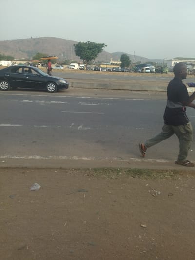 The image shows a man walking on the sidewalk of a paved road in Maraba, Nigeria (latitude: 9.024, longitude: 7.573). He is wearing a black shirt, khaki pants, and brown sandals. He is walking to the right side of the frame. Behind him, a black sedan is stopped in the middle of the road. There are other cars parked on the road, including white vans, a blue car, and a black car. The sidewalk is dusty and covered in dirt. There are a few pieces of trash on the ground. There is a hill in the background. The sky is cloudy and overcast.
