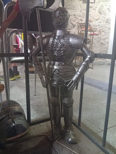 The image depicts a full-body suit of armor, likely a mannequin, displayed behind a metal fence in what appears to be an indoor setting. The armor is made of dark, metallic material and is intricately detailed, featuring a helmet with eye slits, a chest plate with a decorative emblem, segmented arm and leg guards, and a gauntlet. The armor is holding a long polearm, possibly an axe or halberd. In the foreground, to the left, a knight's helmet is placed on the ground. In the background, a stone wall is visible, with a shelf holding several candles. The floor is tiled. Part of a person's arm, wearing a pink sleeve, is visible in the upper left corner, suggesting the presence of a visitor or attendant. The location is identified as Sobótka, Poland.