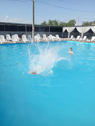 The image captures a sunny day at an outdoor swimming pool. A person is mid-jump into the pool, creating a large splash of water. In the background, a woman with her hair in a bun is swimming. Several white lounge chairs are arranged around the pool, some occupied by people. A metal pole with a sign on it stands near the pool's edge. The area is shaded by a structure with a dark roof and white curtains. Trees and buildings are visible in the distance, suggesting a warm, clear day. The overall mood is one of leisure and recreation. There is no visible text in the image.