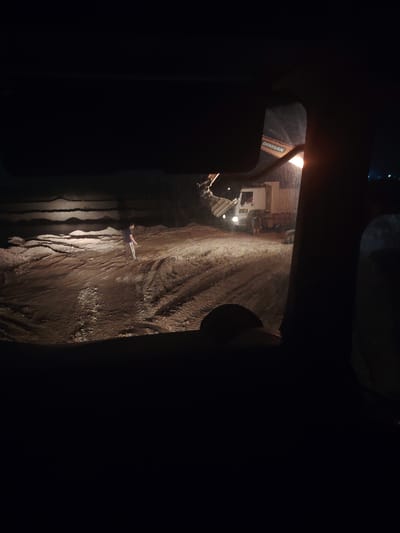 The image depicts a construction site at night in Dacha, Uzbekistan. In the foreground, a dark interior of a vehicle is visible, suggesting the photo was taken from inside. In the middle ground, a person in a dark t-shirt and light-colored pants stands on muddy ground with tire tracks. Behind the person, a white dump truck is parked, and an excavator with "DOOSAN" branding is operating, its light illuminating the scene. The environment appears to be a quarry or excavation area, with layered dirt or sediment visible in the background. The lighting suggests it is dark outside, with artificial light from the construction equipment.