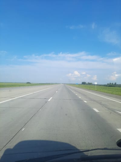 The image depicts a wide, straight asphalt road stretching into the distance under a bright blue sky with scattered white clouds. The road is divided by white lane markings. To the left and right of the road are flat, green fields, with a fence line visible in the distance on the right. There are a few scattered trees and some distant buildings or structures. On the right side of the road, a round white sign with a blue symbol and a smaller white sign with a blue symbol are visible. A dark shadow, likely from a vehicle, is cast across the bottom foreground of the image. The scene appears to be located on the outskirts of Pokrovka, Russia, given the flat terrain, open fields, and the style of road markings and signage.