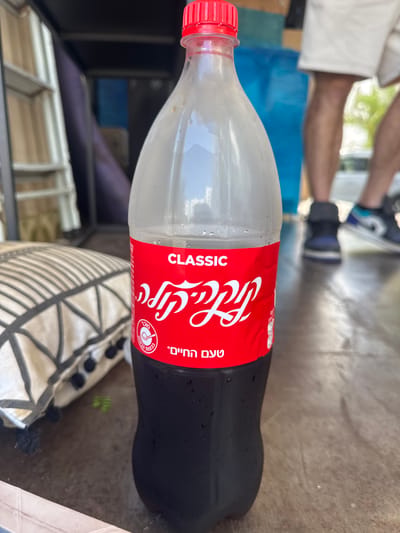A Coca-Cola Classic bottle is positioned in the foreground, its red label partially visible with white lettering. The bottle contains a dark liquid and appears to be sweating, indicating it is cold. In the background, a person is standing, wearing white shorts and sneakers. Other blurred objects and individuals are also present in the background, suggesting an outdoor setting. The ground beneath the bottle is a brown, textured surface. A patterned pillow is visible to the left of the bottle. The lighting suggests it is daytime. The scene is set in Tirat Karmel, Israel.