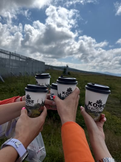 In T/B Seminskii Pereval, Russia, four hands are holding coffee cups in a toast. The cups are white with black lids and feature a design of trees and a deer, with text that appears to read "КОФЕ на высоте 1717" which translates to "Coffee at an altitude of 1717". The background shows a grassy hillside with a metal fence and a cloudy sky. The people are wearing casual clothing, including an orange shirt and a watch with a light purple band.