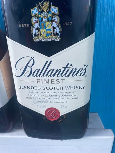 This is a close-up, eye-level, outdoor, medium shot of a bottle of Ballantine's Finest Blended Scotch Whisky. The bottle is dark, with a prominent white label featuring the Ballantine's logo and text. The label states "Ballantine's Finest Blended Scotch Whisky" and provides details about its blending and bottling in Scotland by George Ballantine and Son, Dumbarton, G82 2SS, Scotland. It also indicates a volume of "1 Litre e" and an alcohol content of "40% vol". The bottle is positioned against a bright blue, textured background, likely a wooden surface, suggesting it is outdoors. The overall scene is brightly lit, with some slight shadows and reflections on the bottle's surface. There are no people or animals visible in the image. The location in Бабовіцкі сельскі Савет, Belarus is not directly discernible from the visual information in the image itself, but the image depicts a product that might be found in a store or as part of a personal collection in that location.