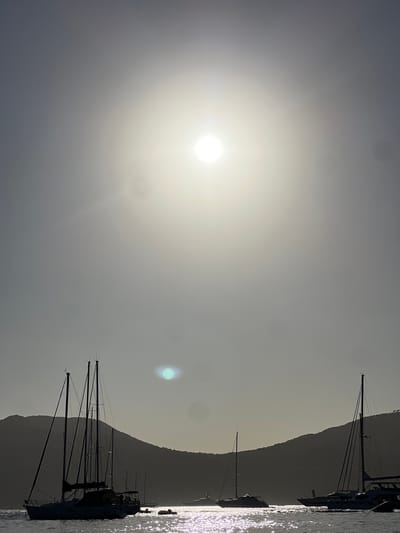The image depicts a serene bay with several sailboats anchored in the sparkling, sun-drenched water. The sun is high in the sky, casting a bright glare and creating a hazy, atmospheric effect. In the foreground, a large sailboat with multiple masts dominates the left side of the frame, its silhouette sharply defined against the luminous sky. Other sailboats of varying sizes are scattered across the bay, their masts and hulls also appearing as dark shapes. In the background, a lush, verdant hillside rises gently from the water, its contours softened by the haze. The water reflects the sunlight in a shimmering pattern, adding to the tranquil ambiance of the scene. The location is identified as the bay of Sant Josep de sa Talaia, Spain, a popular coastal town known for its beautiful beaches and picturesque harbors.