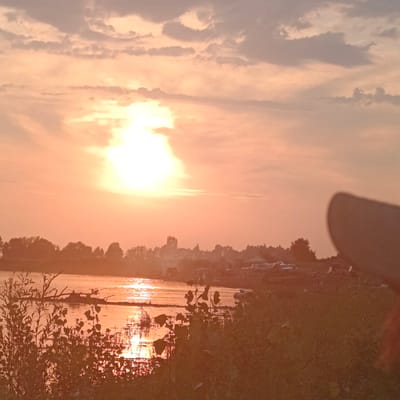 The image depicts a sunset over a body of water, likely a river, in the vicinity of Старый Буртюк, Russia. The sky is filled with clouds illuminated by the setting sun, creating a warm, orange and yellow glow. The sun itself is a bright, diffused light source. On the horizon, a line of trees forms a silhouette against the sky. In the foreground, dense vegetation lines the bank of the water. The water reflects the colors of the sunset, with shimmering light on its surface. There are some indistinct objects in the water, possibly debris or small boats. On the far bank, beyond the trees, there are some structures and what appear to be parked vehicles, suggesting a populated area or industrial site. A blurry, dark object, possibly a hand or a hat, is partially visible in the lower right corner of the frame, obscuring a portion of the scene.