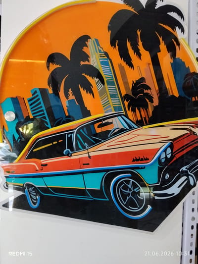This is a digitally created image or artwork, likely a stylized representation of a classic car against a city skyline with palm trees. The dominant colors are orange, teal, and black, creating a vibrant, retro aesthetic. A vintage-style car, painted in stripes of orange and teal with white accents, is prominently featured in the foreground, positioned on a black surface. In the background, a silhouette of a city skyline with tall buildings is visible, interspersed with dark silhouettes of palm trees. The sky is a bright orange, suggesting a sunset or sunrise. The overall scene evokes a tropical, possibly Miami-inspired, vibe. The artwork appears to be mounted on a white background, and there's a silver circular element on the left side, possibly a mounting point or part of the artwork itself. The image also displays a timestamp in the bottom right corner: "21.06.2026 10:31", and the text "REDMI 15" in the bottom left corner, indicating it was captured or created with a Redmi 15 device. The location context provided is Yovon, Tajikistan, which doesn't directly align with the imagery itself, suggesting the artwork might be displayed or created in that location. There are no people visible in the image.