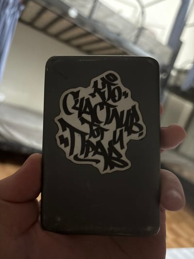 A human right hand holds a dark, rectangular electronic device, possibly a smartphone or power bank, in a vertical orientation. The device exhibits minor scratches and scuffs on its dark grey surface. Affixed to the center of the device's visible surface is an irregularly shaped sticker with a white background and black graffiti-style text in Cyrillic script, reading "НЕ СЧАСТЬЕ ОТ ПАРЫ." The fingers of the right hand grip the device from the sides and bottom. The background is out of focus, revealing indistinct structural elements consistent with an indoor setting, including a blurry metal frame, possibly from a bunk bed or shelving unit, in the upper portion, and a lighter-colored wall or ceiling and dark floor or furniture in the lower portions. No visual information within the image identifies a specific location in Phong Nha Commune, Vietnam.