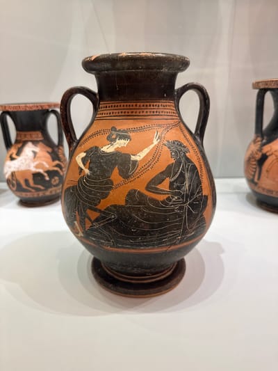 The image shows a reddish-orange ceramic amphora with black painted figures. The amphora has two handles and a narrow neck, widening to a bulbous body that tapers to a footed base. The main decoration on the front of the amphora depicts two figures in black silhouette style. On the left, a standing female figure with her hair in a bun extends her left arm towards a seated male figure on the right. She wears a garment that leaves her right arm bare and a patterned skirt. The seated male figure is bearded and wears a headdress and a draped garment. He holds an object, possibly a lyre, in his lap. Both figures have detailed facial features rendered in thin black lines. The amphora is displayed on a white surface, likely a pedestal in a museum exhibit. To the left and right of the central amphora, other similar ceramic vessels are partially visible, suggesting a collection or display of ancient pottery. The background is a plain, light gray. The location is not discernible from the image alone, but such artifacts are commonly found in museums such as the Archaeological Museum of Heraklion, Greece.