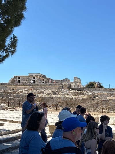 The image depicts a group of tourists exploring the ruins of the Palace of Knossos, an archaeological site located in the Heraklion Municipal Unit, Greece. The ruins, constructed from stone blocks, feature remnants of walls, pillars, and paved pathways. Several people, dressed in casual summer attire, are visible within the scene, some appearing to be part of a guided tour. One individual in the foreground, wearing a blue t-shirt and a blue baseball cap, is looking to the left. Another person, in a blue t-shirt and a black baseball cap, is in the middle ground, holding what appears to be a small object. The background reveals the expansive, multi-level structure of the palace complex under a clear blue sky. Vegetation, including the branches of a pine tree in the upper left corner, is also present. The overall atmosphere suggests a sunny, daytime visit to a historical location.