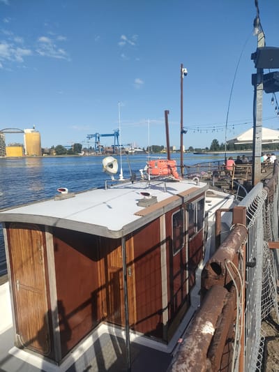 This is a photograph taken from the deck of a boat docked at an outdoor seating area. In the foreground, the side of the boat, constructed with reddish-brown wood paneling and white accents, is visible. The boat has a small cabin with wooden doors and windows. A white railing with a security fence runs along the right side of the image, separating the deck from a sandy area.

Beyond the boat, a body of water is visible, reflecting the blue sky with scattered white clouds. In the distance, across the water, there are industrial structures, including large yellow silos and blue cranes, indicative of a port or industrial zone. A bridge can also be seen in the background.

The outdoor seating area appears to be a restaurant or bar, with picnic tables and a white canopy structure. Several people are seated at the tables, enjoying the outdoors. The overall scene suggests a sunny day in a waterfront area.

The industrial structures and port activity in the background, particularly the large cranes and silos, are characteristic of the Port of Gdańsk. The presence of a boat docked at a waterside venue further supports this location. Therefore, this scene is likely situated in Gdańsk, Poland, along the waterfront, possibly near the port facilities.