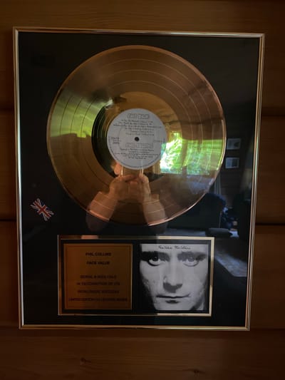 This media file shows a framed gold record award for Phil Collins' album "Face Value". The award is mounted on a black matte background within a silver-toned frame. The gold record itself is prominently displayed, with its grooves reflecting light. The center label of the record includes text such as "Face Value", "Phil Collins", and a list of song titles and their durations. To the right of the record, there is a black and white photograph of Phil Collins, with his name and "Face Value" written vertically next to it. Below the photograph is a gold plaque with text detailing the award, including "PHIL COLLINS", "FACE VALUE", "SERIAL # RGG-134-2", "IN RECOGNITION OF ITS WORLDWIDE SUCCESS", and "LIMITED EDITION COLLECTORS SERIES". A small Union Jack flag is also visible in the lower right corner of the matte. The overall presentation suggests a celebratory or commemorative item related to the success of the album. The lighting suggests it is daytime, with reflections of what appears to be greenery and possibly an interior space visible on the gold record.