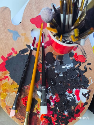 A top-down, close-up view showcases an artist's wooden palette covered in various splotches and smears of paint in red, black, white, and yellow. Three paintbrushes are positioned on the palette: one black-handled brush with a pink tip, a yellow-handled brush, and another black-handled brush with a white tip. A red cup filled with numerous paintbrushes is placed at the top right of the palette. The environment is an indoor setting, likely an art studio, but its specific location in Melhus, Norway, cannot be determined from the image alone. There are no people or interactions depicted in the scene.