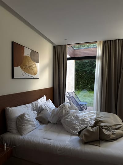 The image depicts a bedroom in Bakovići, Montenegro. The bed is unmade, with white sheets and a duvet thrown back. Several pillows are scattered on the bed. A piece of abstract art hangs on the wall above the headboard. To the right, a large window with curtains leads to an outdoor patio with two chairs and a small table, overlooking a grassy area surrounded by trees. The lighting suggests it is daytime, possibly late afternoon or early morning, with sunlight casting shadows.