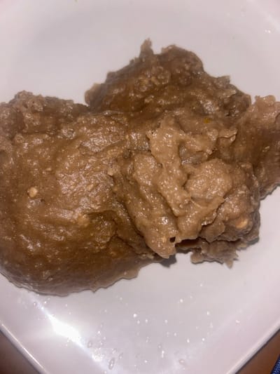 A mound of brown, slightly textured, cooked food rests on a white plate. The food appears dense and somewhat lumpy, with subtle variations in color suggesting varied ingredients or cooking processes. Moisture is visible on the surface of the food and on the plate. The plate is clean, with a few scattered water droplets near the food.