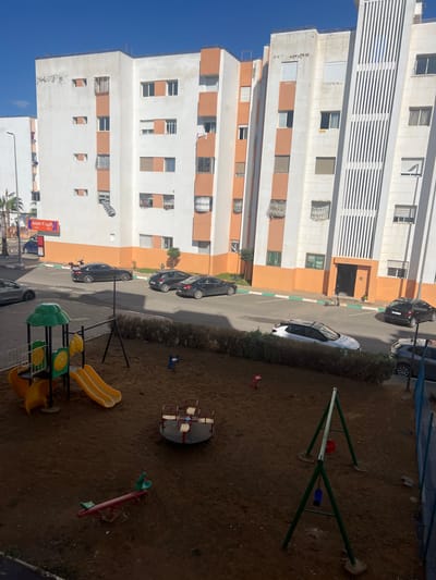 The image depicts an outdoor scene in a residential area of Skhirat, Morocco, under a clear blue sky. In the foreground, a children's playground features brown dirt ground, partially shaded. Equipment includes a green and yellow play structure with two yellow slides, a red and white merry-go-round, a green metal swing set with two empty swings (one red, one blue), a red seesaw, and two empty animal-shaped spring riders (one blue, one pink).
A low hedge separates the playground from a paved street or parking area in the midground. Several vehicles are parked: two dark sedans are parked parallel on the left, a white SUV with a dark roof is parked parallel behind the hedge, and two dark vehicles (one sedan, one SUV) are parked diagonally on the right. Parking spaces are delineated by green and white lines on the asphalt. A solitary, dark-clad individual is visible near the entrance of the rightmost building.
The background is dominated by multi-story residential buildings, primarily white with orange horizontal accent panels around windows and balconies. Many windows feature security bars, decorative grilles, or closed white roller shutters. The building on the far right exhibits distinctive vertical white striped paneling on its facade. On the ground floor of the leftmost building, a commercial establishment with a red awning and "Marjane Express" signage is visible. A street light pole and palm trees are present on the far left. The scene is brightly lit by natural sunlight, with shadows indicating the sun's position.