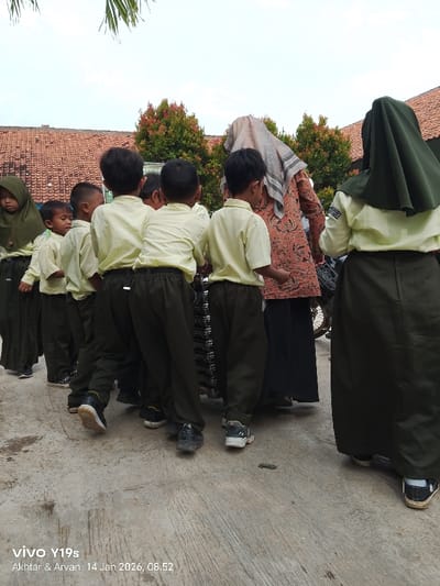 A group of young children in light yellow shirts and dark green pants line up, facing away from the camera. Most have short hair. They appear to be holding onto a dark object with multiple slats. Two women in headscarves and long skirts stand with them, one in green and the other in a printed fabric. The group stands on a concrete surface in an outdoor setting, with buildings and greenery visible in the background. A motorcycle is parked nearby.