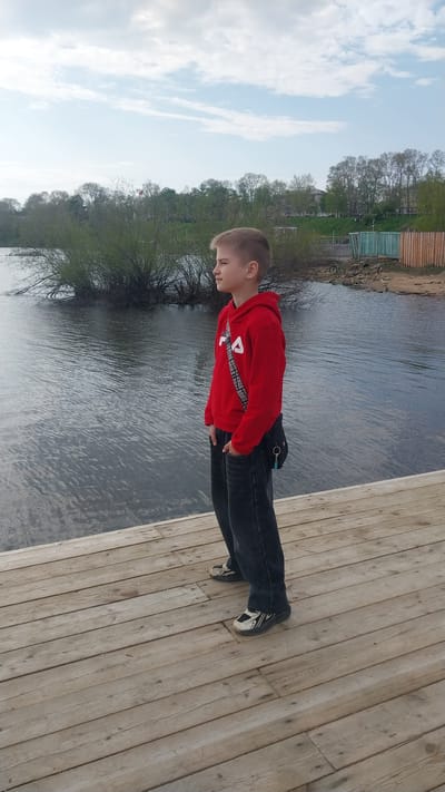 The image displays a young boy standing in profile on a wooden pier, with a large body of water to his left, consistent with a riverside location in Nikolayevsk-on-Amur, Russia. The boy, positioned slightly right of the frame's vertical center, faces left with his eyes either closed or cast downwards. He is dressed in a red hooded sweatshirt featuring a white "FILA" logo on the chest, dark grey jeans, and athletic shoes that are predominantly white/cream with black and grey accents. A black shoulder bag with a white, text-printed strap is worn across his chest, and his hands are placed inside his jeans pockets.

The pier is constructed from light-colored, horizontally laid wooden planks. To the boy's left and extending into the midground, the water surface exhibits minor ripples. Beyond the water, dense green and brown shrubbery occupies an area that could be an island or the opposite riverbank. In the far background on the left, deciduous trees with emerging foliage are visible, along with a distant red flag atop a pole. To the right side of the frame, a sandy bank abuts the water, beside which stands a structure made of vertical wooden planks; some planks are painted teal-green, while others are unpainted. A bicycle is present on the ground near this structure. The distant background reveals additional trees and architectural forms suggestive of a town, all beneath a sky characterized by scattered white clouds and blue patches.