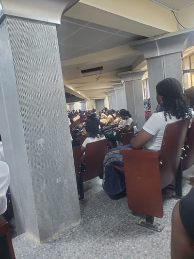 In Kason, Nigeria, a dimly lit indoor space, possibly a church or conference hall, accommodates a gathering of people seated in rows of brown chairs. The hall features a perforated metal pillar in the foreground, a suspended acoustic tile ceiling, and decorative architectural elements. A woman wearing a white top and dark headscarf is seated in the front row, facing away from the camera. Other individuals, mostly women dressed in modest attire, are visible throughout the hall, some looking ahead, others engaged with their phones or conversing. The overall atmosphere appears calm and attentive.
