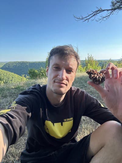 The image is a selfie taken outdoors, featuring a young man with short brown hair and light skin. He is wearing a black long-sleeved shirt with a large yellow graphic and the text "THE ORIGINAL" on the sleeve, and black shorts. He is sitting outdoors on dry grass, with his right hand raised, holding a pinecone. In the background, there is a lush, green landscape with rolling hills covered in trees, under a clear blue sky. The location is described as the city of Nihyn, Ukraine. The lighting suggests it is daytime, likely late afternoon due to the warm tones.
