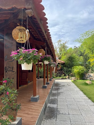 The photograph shows a view of a covered walkway in Liên Nghĩa, Vietnam. The walkway is lined with wooden posts supporting the roof, and each post has a hanging flower pot and/or wind chime. The roof has a traditional red tile design. The walkway is paved with patterned grey tiles, and a green grassy area with trees is visible on the right. The building's architecture is traditional, with decorative wooden elements and a red tile roof. Hanging baskets with purple and green plants adorn the beams. A path leads away from the walkway.