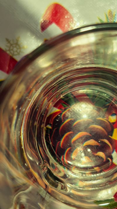 A top-down close-up shot captures the interior of a clear glass, revealing a colorful pattern beneath it. The pattern appears to be a stylized Christmas design, featuring elements such as a snowflake, red and green ribbons, and a pinecone. The glass's ribbed texture refracts the light and the colors of the pattern, creating a kaleidoscopic effect. The lighting suggests a bright, possibly sunny environment. The scene is devoid of people or any discernible action. The location is identified as Troițcoe, Moldova.