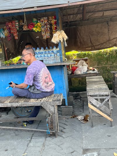 An elderly Asian man with glasses and a mustache sits cross-legged on a wooden bench in front of a blue food stall. He wears a purple tie-dye t-shirt with a pink Vespa scooter graphic on the back and dark pants. He holds a black smartphone and a cigarette in his left hand. A gray fanny pack is worn around his waist. Behind him, on the stall counter, are rows of bottled water. The stall has various snacks hanging from the front. The ground in front of the stall is paved with cracked concrete. A wooden cart wheel is visible beneath the bench. A lush green vine-covered wall is visible in the background. This scene is located in Gedang, Indonesia.