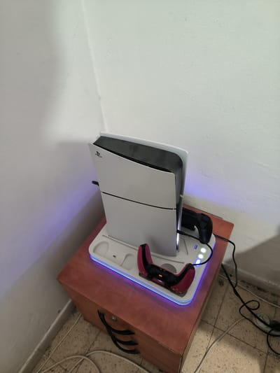 In a room with off-white walls and tiled flooring, a wooden cabinet stands in the corner. On top of the cabinet, a white PlayStation 5 console is positioned vertically on a white charging stand. The stand has blue LED lighting around its base and features two controller charging docks. One dock holds a black controller, while the other holds a red and black controller. The scene is lit by overhead lighting, casting soft shadows. This image was captured in Be'er-Sheva, Israel.