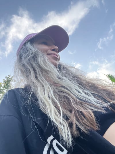A low-angle, outdoor, close-up selfie captures a woman wearing a purple baseball cap and a black t-shirt. Her long, blonde and brown hair is blown to the side by the wind, obscuring part of her face. She is smiling and looking off to the right, with a bright blue sky and scattered white clouds in the background. The image appears to have been taken during the day, as indicated by the natural lighting. The background also shows hints of greenery, suggesting a natural or park-like setting.