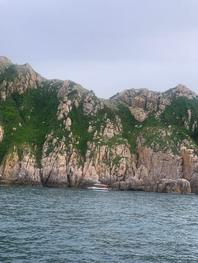 The image shows a rocky coastline with sparse green vegetation on a hazy day. The sea in the foreground is a deep blue with visible ripples. A white boat with orange accents is anchored near the shore, which is lined with weathered, light-colored rocks. The sky is overcast and a pale gray. The scene appears to be the coastline of Vinh Hai, Vietnam, known for its dramatic rock formations and clear waters.