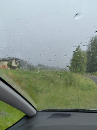 The media file depicts a view through a car's windshield on a rainy day. The windshield is covered with numerous water droplets, distorting the scene beyond. The background shows a rural landscape in Belarus, specifically within the Garanski rural council. A light-colored building, possibly a house, with a dark roof is visible on the left side of the frame. Adjacent to it is a lush green field or meadow. Further into the distance, several trees with dark green foliage are present, and a narrow, dark-colored road or path can be seen on the right. The sky is overcast and appears grey, indicating a cloudy and wet weather condition. The overall mood conveyed is subdued and somber, consistent with rainy weather. The shot is taken from inside a vehicle, with a portion of the car's interior, specifically a light grey trim piece and a dark dashboard element, visible in the foreground. No people are visible in the image. The time of day is indeterminate due to the overcast conditions. No text is visible in the image.