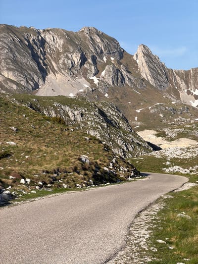 The image depicts a narrow asphalt road curving along the side of a mountain in Boričje, Montenegro. To the left of the road, a steep, rocky mountainside rises with patches of grass and lingering snow. The sky is visible in the upper left, a clear blue. The road surface is gray and appears to be well-maintained. To the right of the road is a sloping terrain covered with grass, rocks, and sparse vegetation. There are no people or animals visible in the frame. The photograph appears to have been taken during the day, with sufficient natural light.