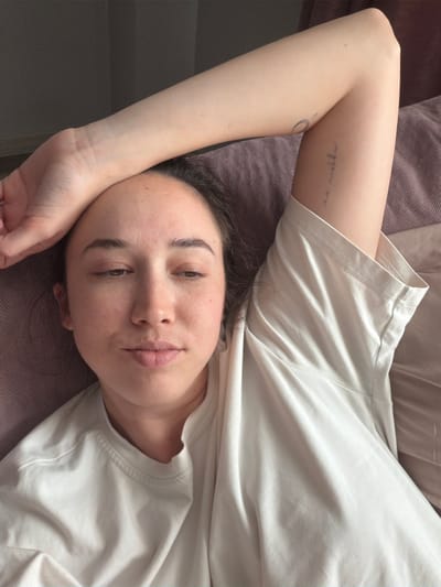 A woman lies on a mauve upholstered surface, likely a sofa or bed. She wears a white t-shirt. Her left arm is raised above her head. Tattoos are visible on her inner forearm. She looks directly at the camera. The location in the city of Novoalexandrovka, Russia cannot be determined from this image.