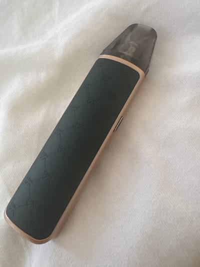 A dark green, elongated vape pen with a gold trim and a translucent dark gray mouthpiece rests diagonally on a textured white fabric surface. The vape pen features a subtle embossed pattern of interconnected diamond shapes. The fabric surface appears to be a cloth or blanket, possibly a bedsheet, with visible texture and folds. The lighting suggests an indoor setting. There are no people or other objects in the immediate vicinity. The image does not depict any location in the city of Schkopau, Germany.