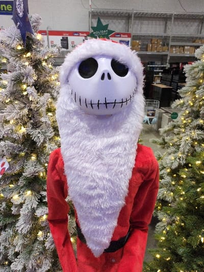 The image features a Jack Skellington Santa Claus figure nestled among snow-flocked Christmas trees adorned with warm white lights. The figure sports a white, fluffy beard and a red Santa suit. The contrast between the eerie character and the festive holiday decor creates a whimsical and unique visual. Shelving and boxes are visible in the background, suggesting a retail setting.