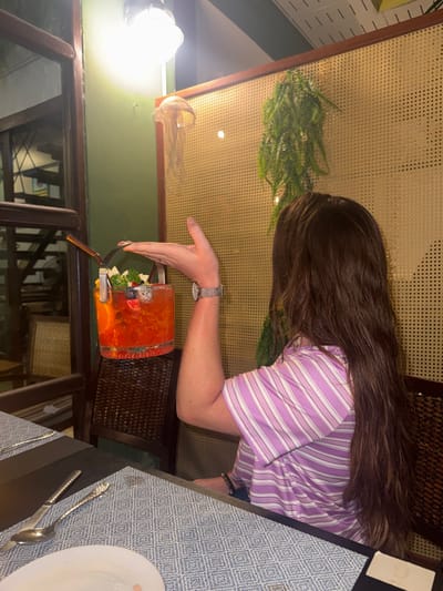An individual, viewed from behind, is seated at a table within a restaurant or bar in Ban Ko Li Pe, Thailand. The person has long, dark brown hair and is wearing a purple and white horizontally striped polo shirt. Their left arm is raised, with the hand open and palm upward, supporting the metal handle of a clear glass bucket-shaped cocktail container. A silver-toned watch is visible on the left wrist. The cocktail contains a reddish-orange liquid with ice, garnished with slices of orange, blueberries, pieces of what appears to be dragon fruit, and green herbs. A metallic straw is inserted into the drink.
The table features a dark surface partially covered by a light blue and white geometric patterned placemat. A silver spoon and knife with textured handles rest on the placemat, alongside a partially visible white plate containing a small, light-colored food item. In the background, a dark brown wicker chair is visible, reflecting the cocktail container.
The establishment's interior features a light green wall on the left, transitioning to a light brown woven rattan screen or partition on the right. A bright, white industrial-style light fixture hangs overhead, illuminating a translucent, light-colored jellyfish-like decorative object affixed to the green wall. A cluster of green foliage, possibly artificial ferns, hangs against the rattan screen. Dark wooden structural elements, potentially window frames or beams, are visible on the far left, with a blurred view of an interior staircase or shelving unit behind them.